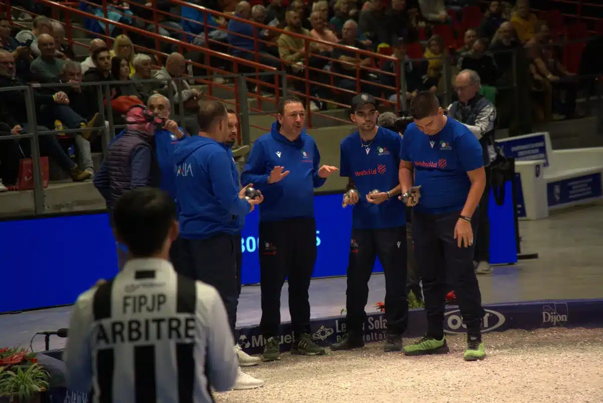 Les règles de la Pétanque : le guide complet et à jour 4 Arbitre de la FIPJP, de dos, face à une équipe qui se concerte boules en main sur un terrain de compétition de Pétanque