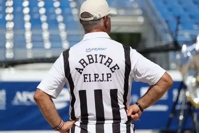 Arbitre de la FIPJP vu de dos, en maillot rayé noir et blanc, sur un terrain de compétition de Pétanque