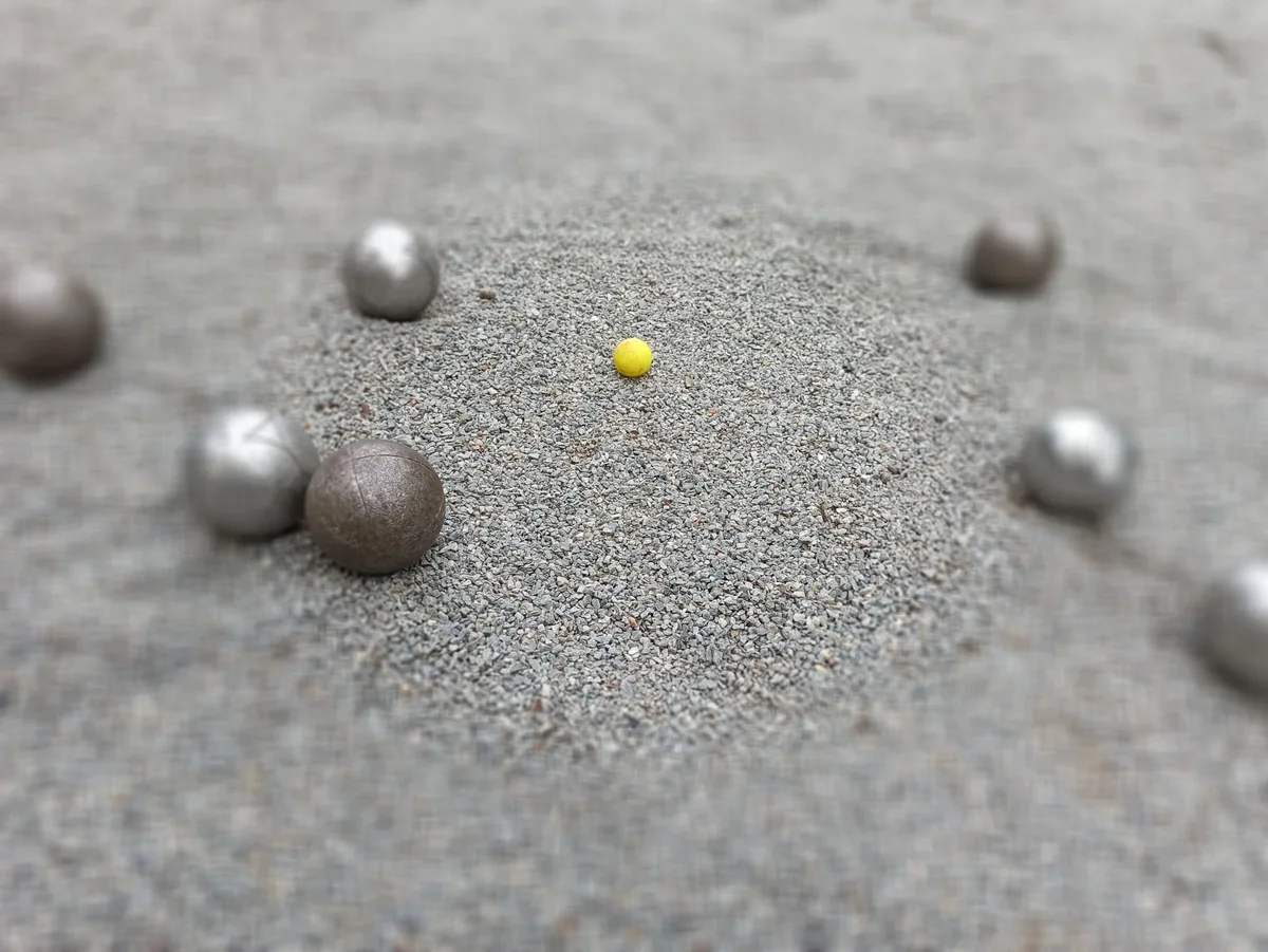 Les règles de la Pétanque : le guide complet et à jour 3 Cochonnet jaune au centre, entouré de boules de Pétanque à des distances différentes, sur un terrain en gravier