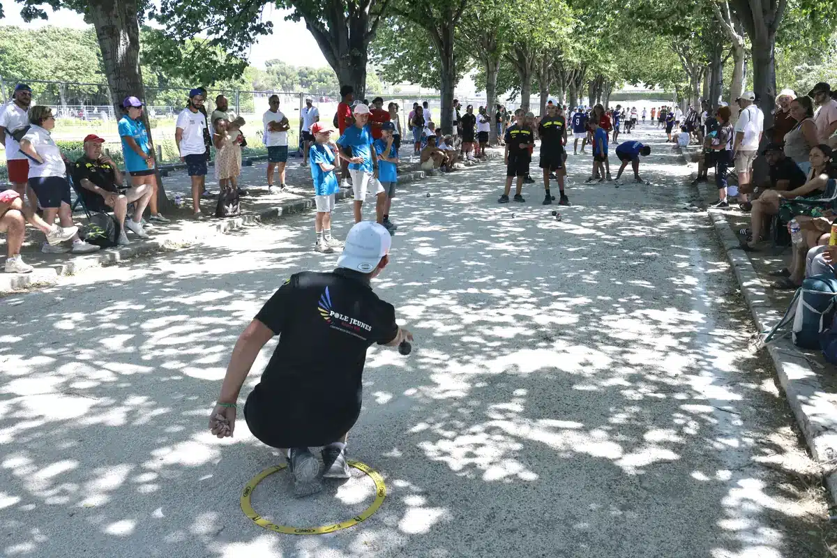 Choisir ses boules de Pétanque : tableau des diamètres, poids et dureté 3 Joueur accroupi dans le cercle, vu de dos, qui s'apprête à pointer sur un terrain ombragé