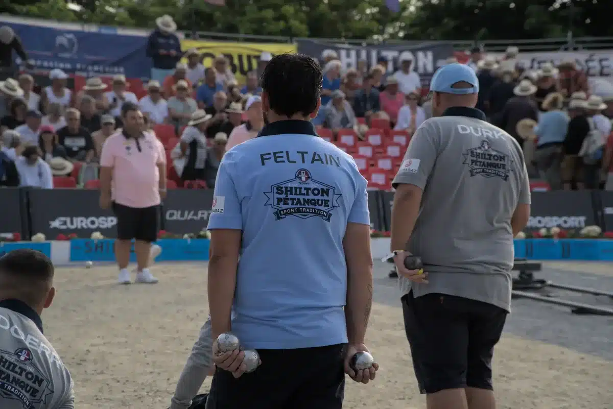Shilton Pétanque : polos, casquette et tenue des Masters, notre guide d'achat 4 Deux joueurs vus de dos, boules en main, en polos Shilton floqués à leur nom, devant des tribunes pleines