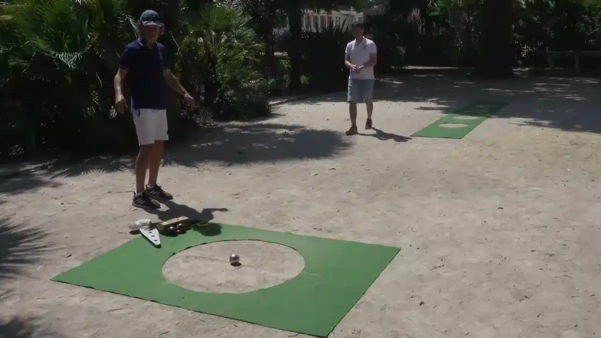 Precision shooting in petanque: rules, 5 workshops and practice 8 Precision shooting area marked out on a shaded terrain: green target circle mat with the target boule in the centre and the template beside it, Jean-François near the target and Alexandre in the distance, by the throwing circle mats